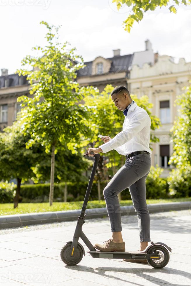 jovem afro-americano usando telefone celular em pé com uma scooter elétrica em uma rua foto
