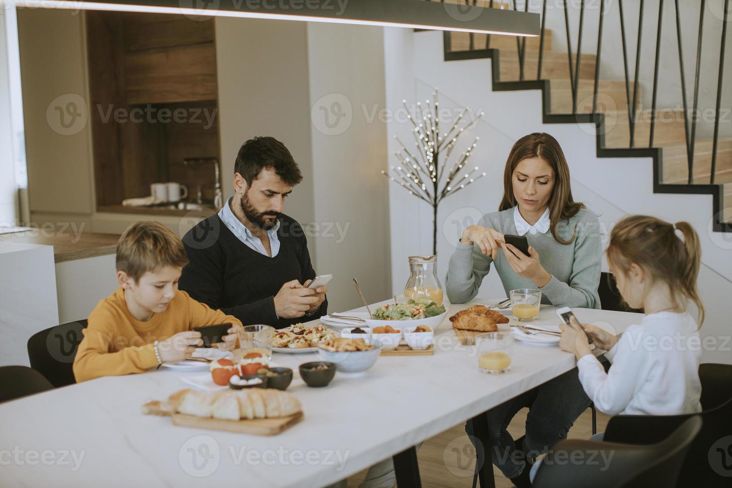 família usando telefones celulares enquanto toma café da manhã na mesa de jantar no apartamento foto