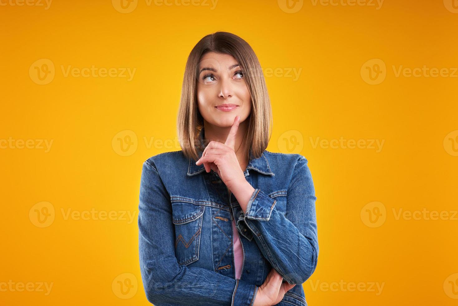 close-up da mulher em jaqueta jeans pensando e apontando sobre fundo amarelo foto