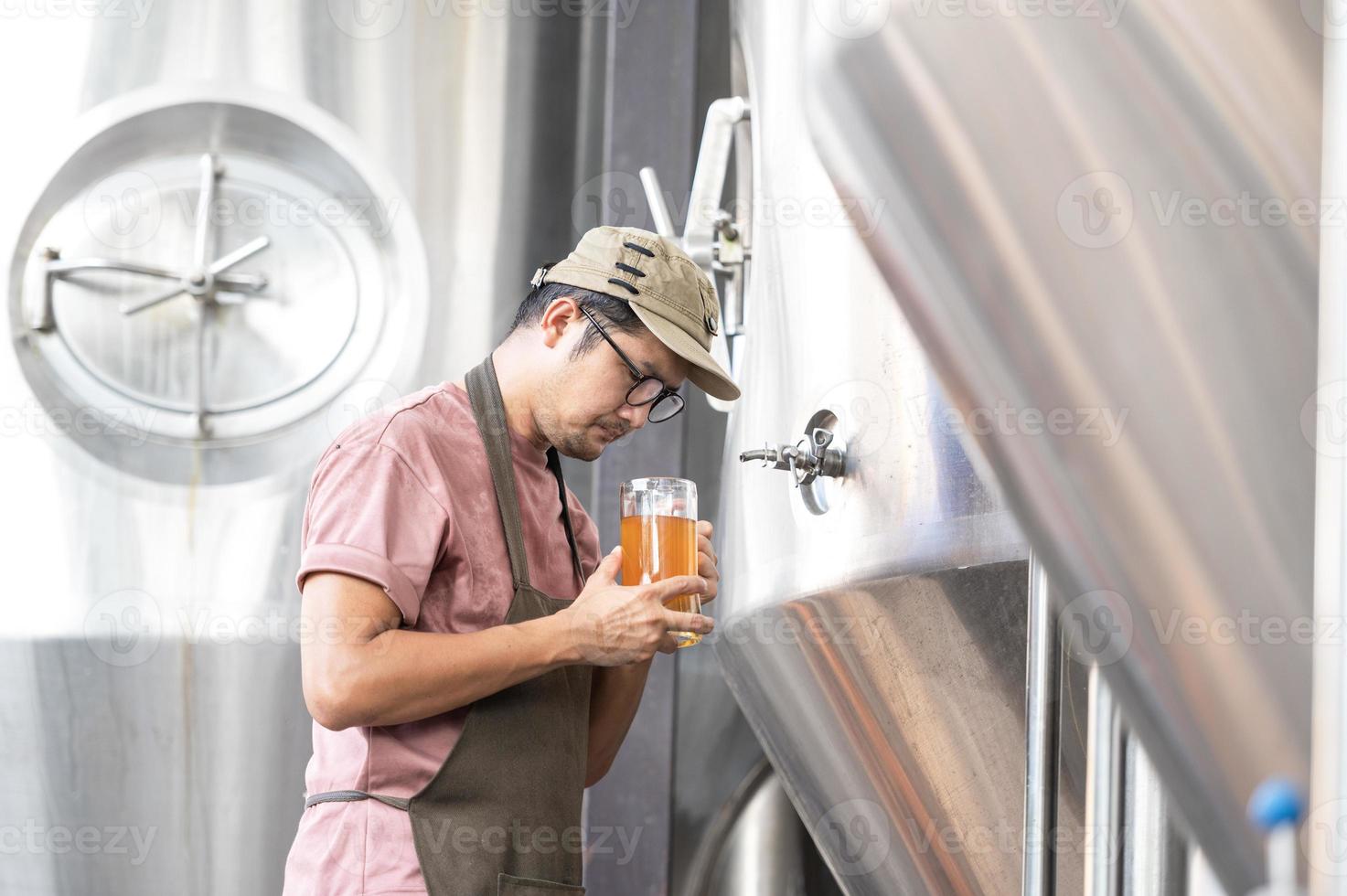 jovem trabalhador asiático inspecionando a qualidade da cervejaria com um copo de cerveja artesanal avaliando a aparência visual após a preparação enquanto trabalhava em uma cervejaria artesanal de processamento. foto