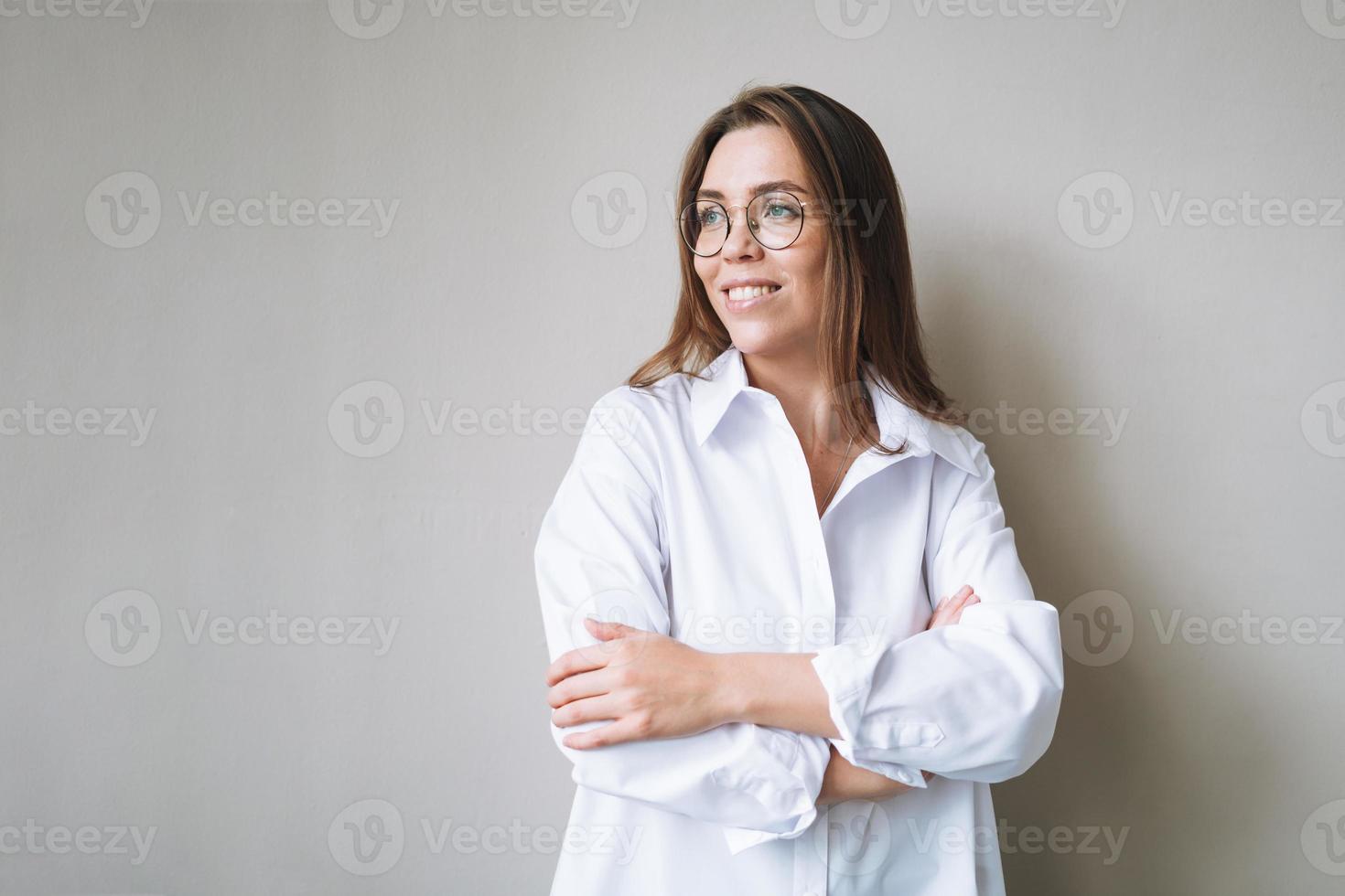 retrato de jovem morena sorridente em óculos e camisa branca perto da parede cinza foto