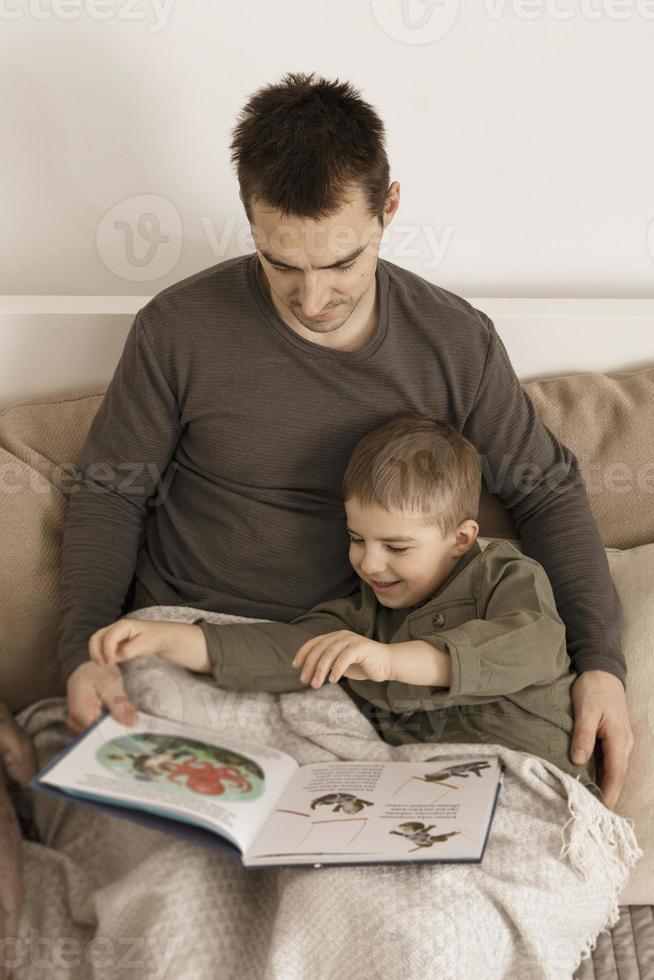 pai e filho lendo um livro na cama em casa. jovem atraente e menino descansando no quarto. cores naturais da terra. ambiente aconchegante. pai lê um conto de fadas para seu filho. foto