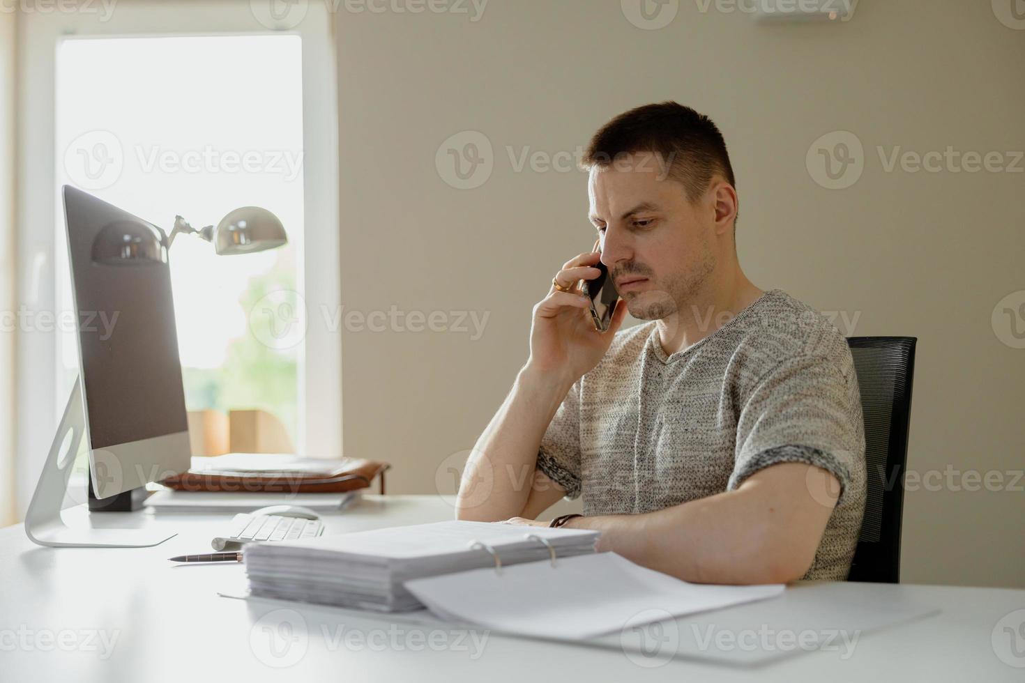 jovem homem caucasiano sentado em sua mesa no escritório, trabalhando com computador e se comunicando por smartphone. jovem empregado tem um dia de trabalho no escritório. empresário fala ao telefone sobre seu negócio foto