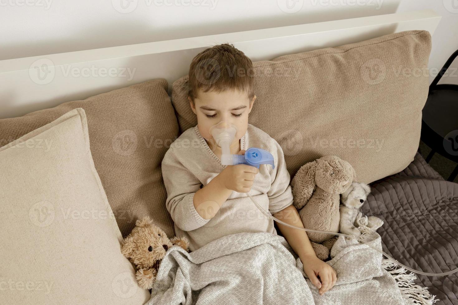 menino doente com inalador para tratamento de tosse. garoto indisposto fazendo inalação na cama dele. temporada de gripe. procedimento médico em casa. interior e roupas em cores naturais da terra. ambiente aconchegante. foto