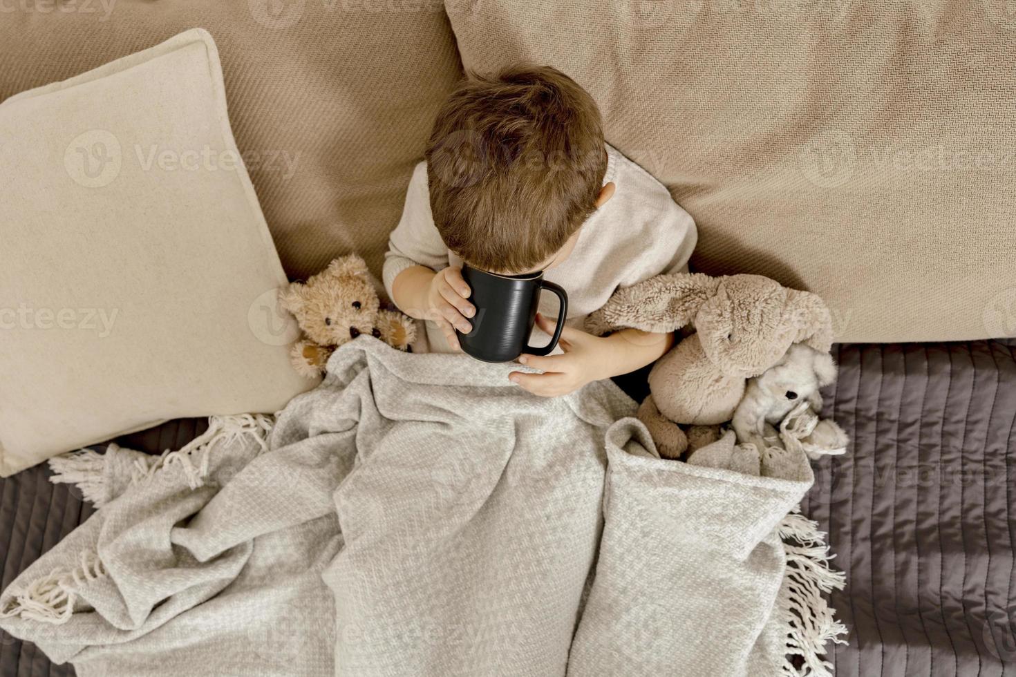 menino doente bebendo chá quente na cama em casa. criança doente, doente enrolada em um cobertor, com caneca em seu quarto. temporada de gripe. interior e roupas em cores naturais da terra. ambiente aconchegante. foto