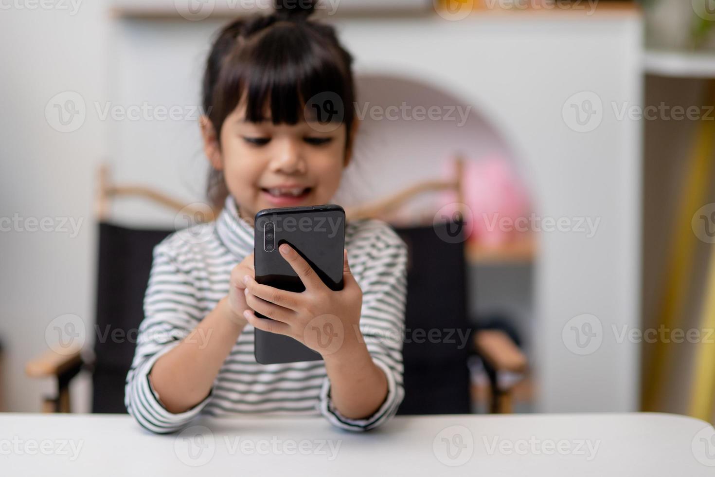menina bonitinha usa smartphone enquanto está sentado no sofá da sala de estar. criança navegando na internet no celular, navega pela internet e assiste desenhos animados online em casa foto