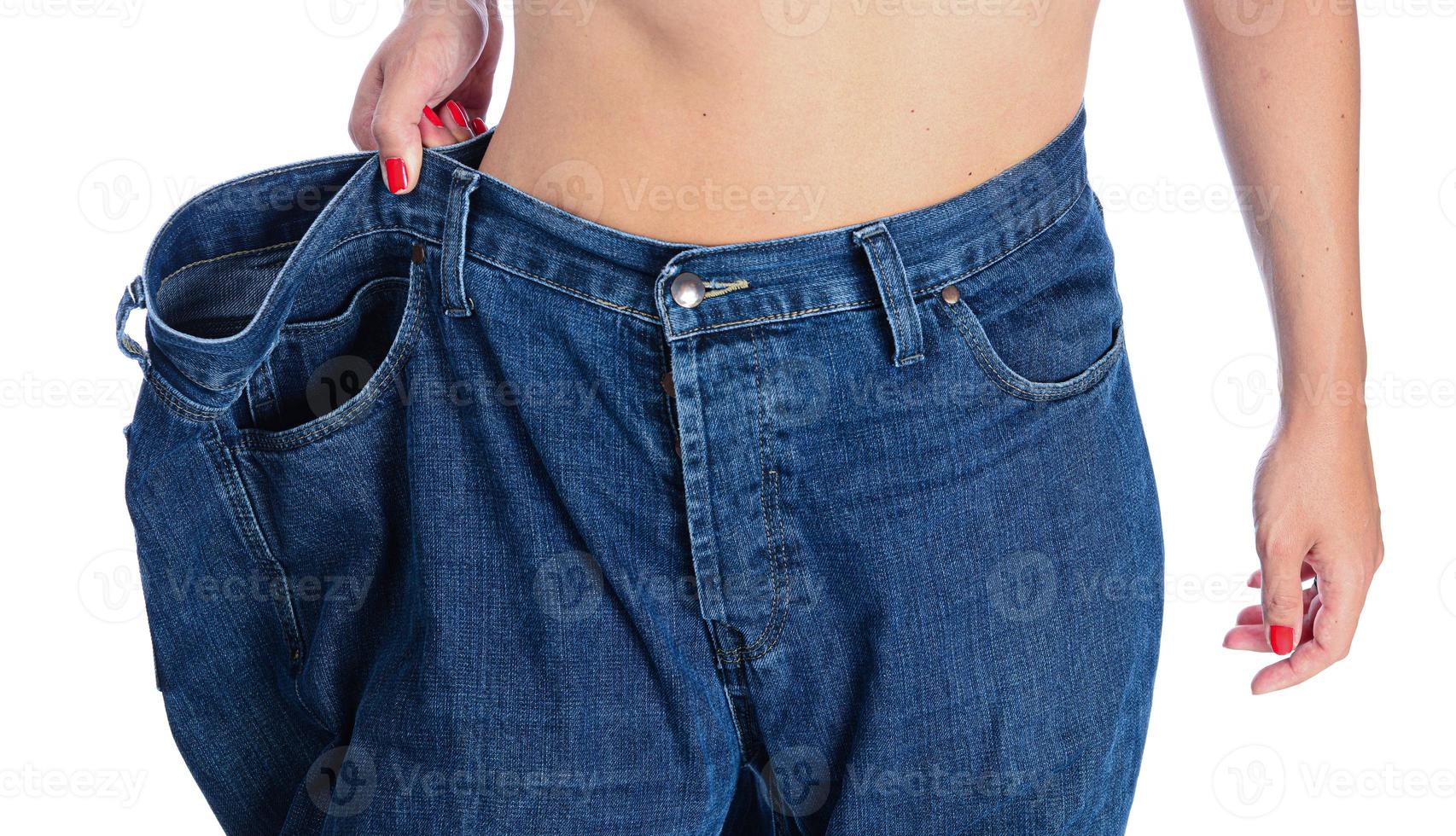 seção intermediária de uma mulher segurando seu jeans azul. menina com jeans grandes isolados no fundo branco. mulher mostra sua perda de peso. conceito de estilo de vida saudável foto