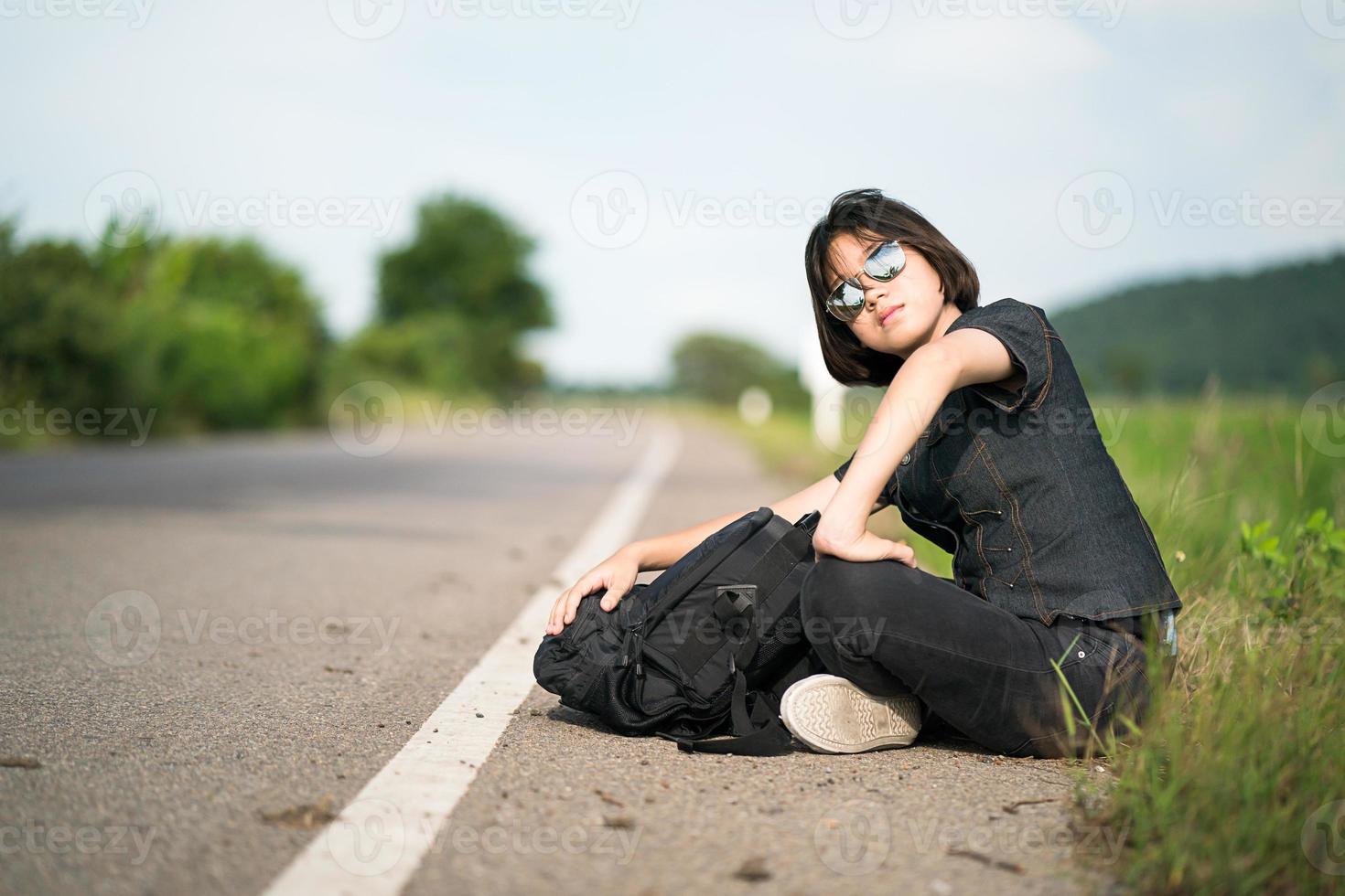 mulher sente-se com mochila pedindo carona ao longo de uma estrada na zona rural foto