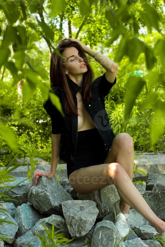 jovem de beleza senta-se em uma rocha em jaqueta preta e calcinha com os olhos fechados foto
