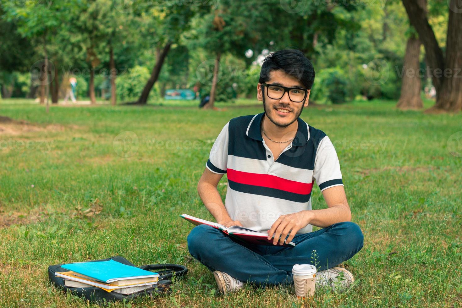 lindo cara sorridente em óculos sentado na grama com livros foto