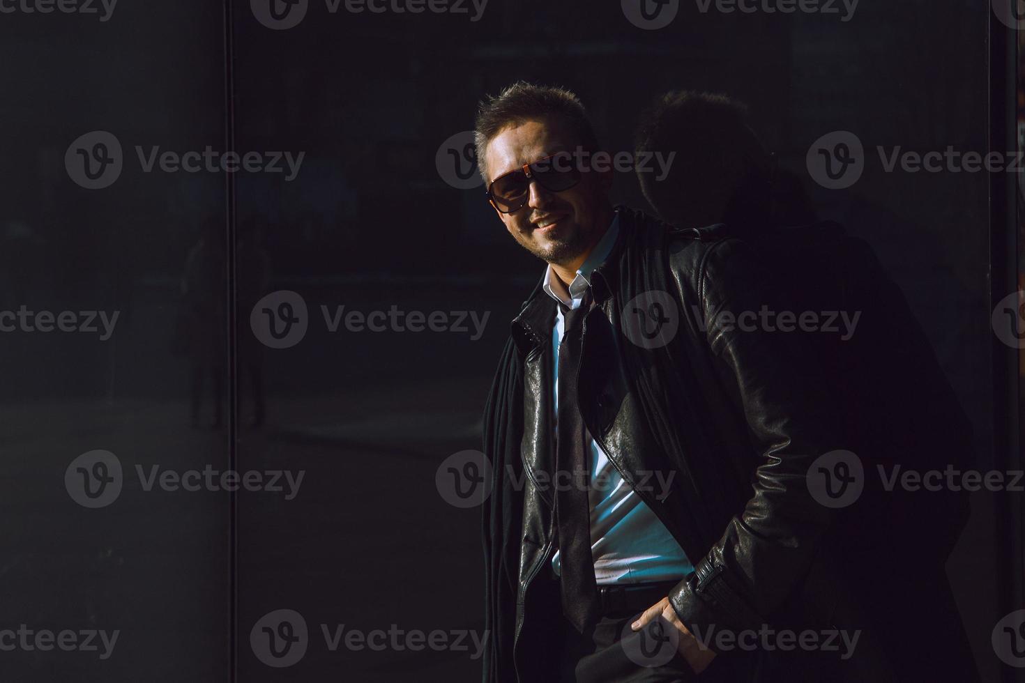 foto escura de homem de negócios divertido em terno de couro e óculos escuros perto da parede preta