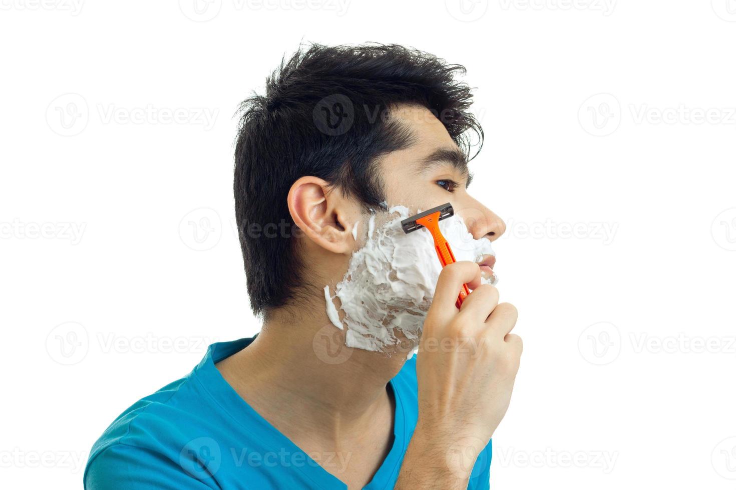 jovem de cabelo preto fica de lado na frente da câmera com a espuma nas bochechas e raspa a barba close-up foto