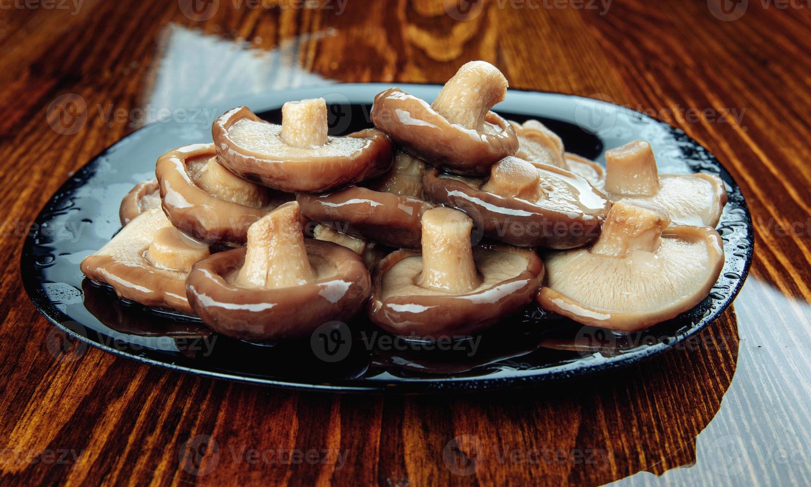 uma pilha de cogumelos em conserva em um prato preto sobre uma mesa de madeira. comida saudável da aldeia. cogumelo de leite preto em conserva. foto