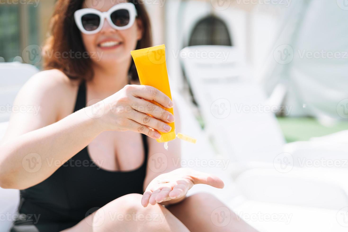 elegante jovem feliz plus size corpo positivo em maiô preto e óculos de sol com creme protetor solar nas mãos na espreguiçadeira perto da piscina, férias de verão foto