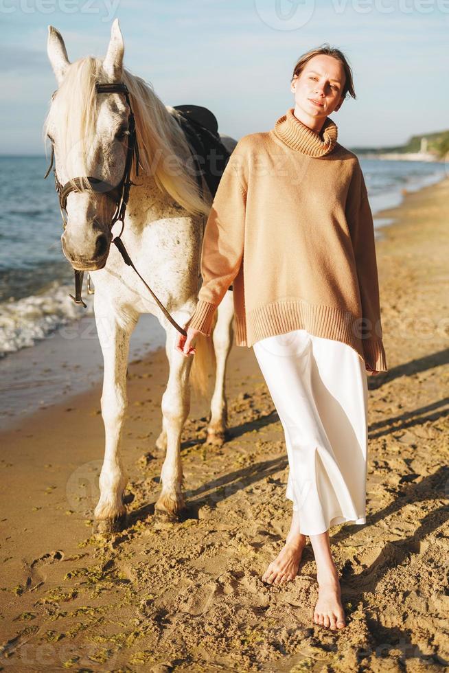 jovem de suéter bege com cavalo branco sobre fundo de paisagem marinha foto