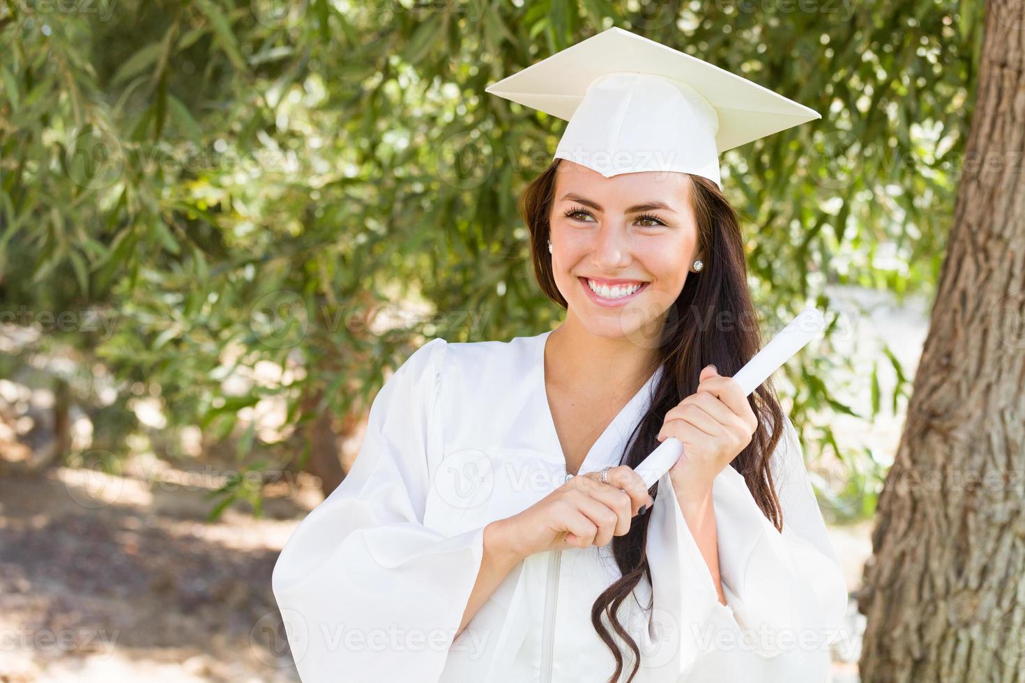 garota atraente de raça mista comemorando a formatura do lado de fora em boné e vestido com diploma na mão foto