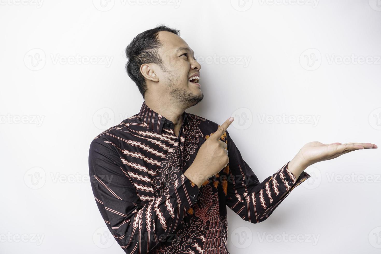 homem asiático animado vestindo camisa batik apontando para o espaço da cópia ao lado dele, isolado pelo fundo branco foto