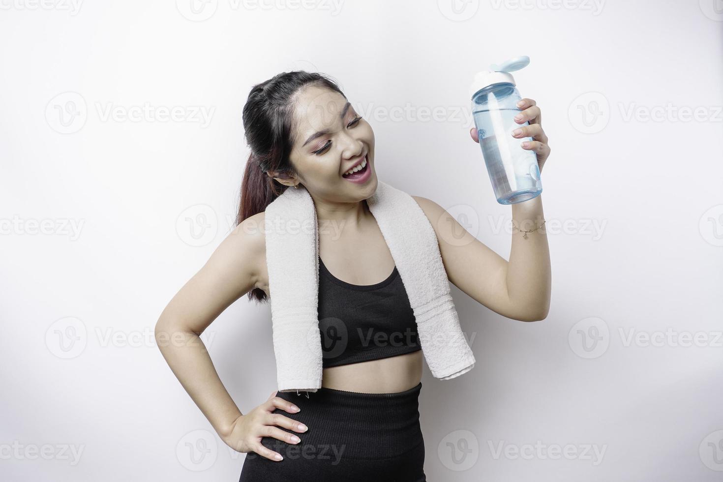 mulher asiática esportiva posando com uma toalha no ombro e segurando uma garrafa de água, sorrindo e relaxando após o treino foto