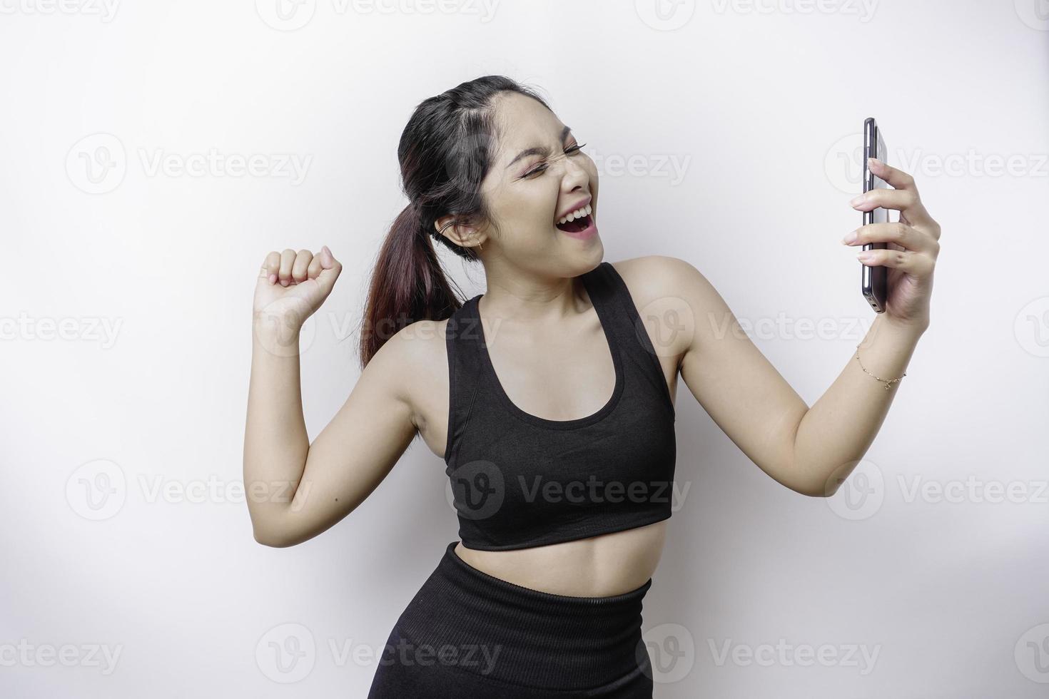 uma jovem mulher asiática desportiva com uma expressão de sucesso feliz vestindo roupas esportivas e segurando o smartphone isolado pelo fundo branco foto