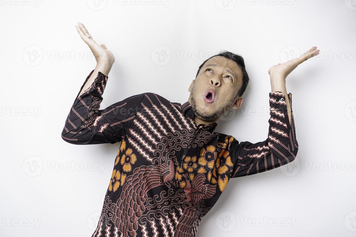 homem asiático chocado vestindo camisa batik apontando para o espaço da cópia em cima dele, isolado pelo fundo branco foto