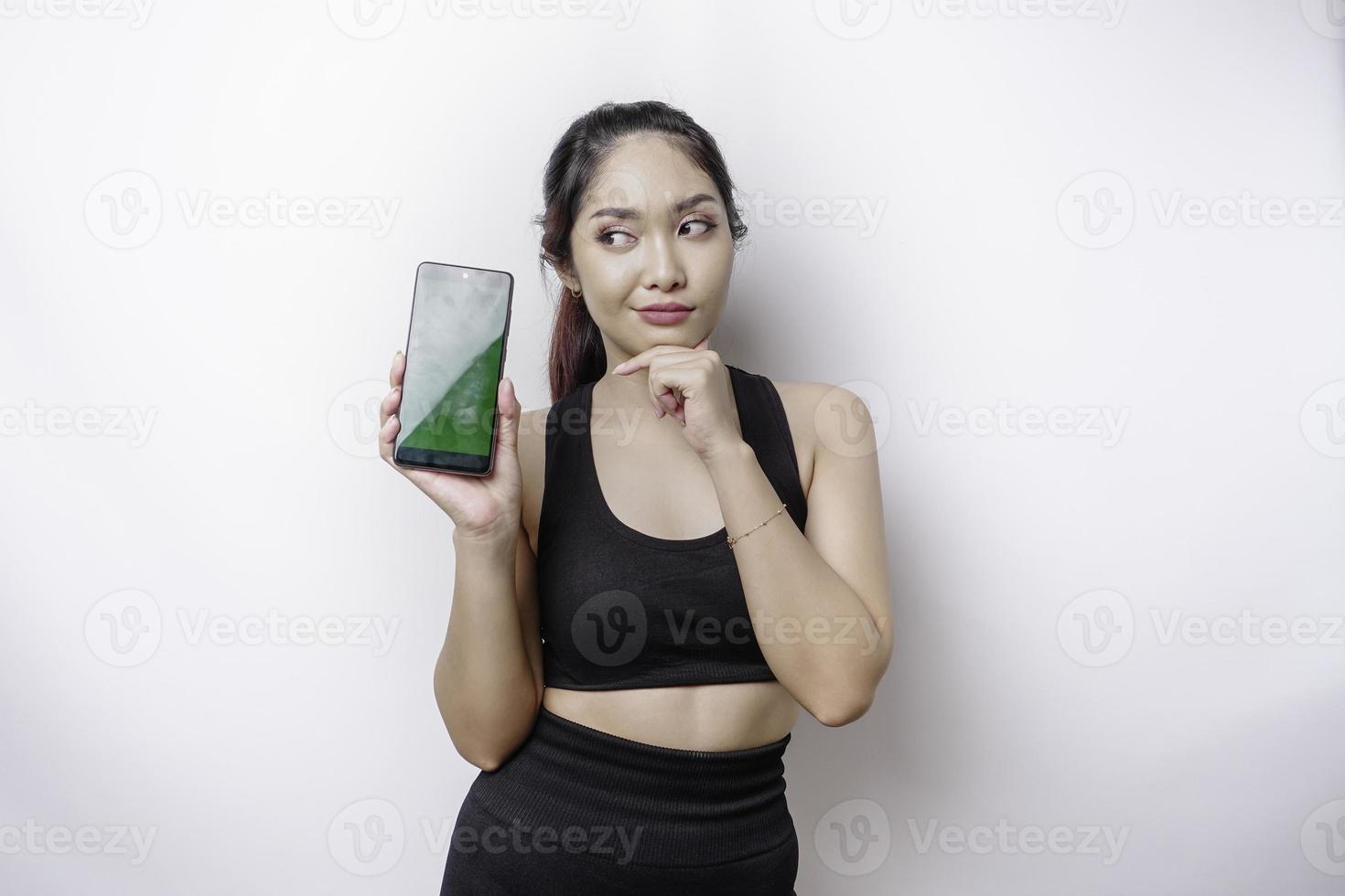 uma jovem esportiva pensativa está vestindo roupas esportivas enquanto segura o queixo e mostra a tela verde em seu telefone, isolada pelo fundo branco foto