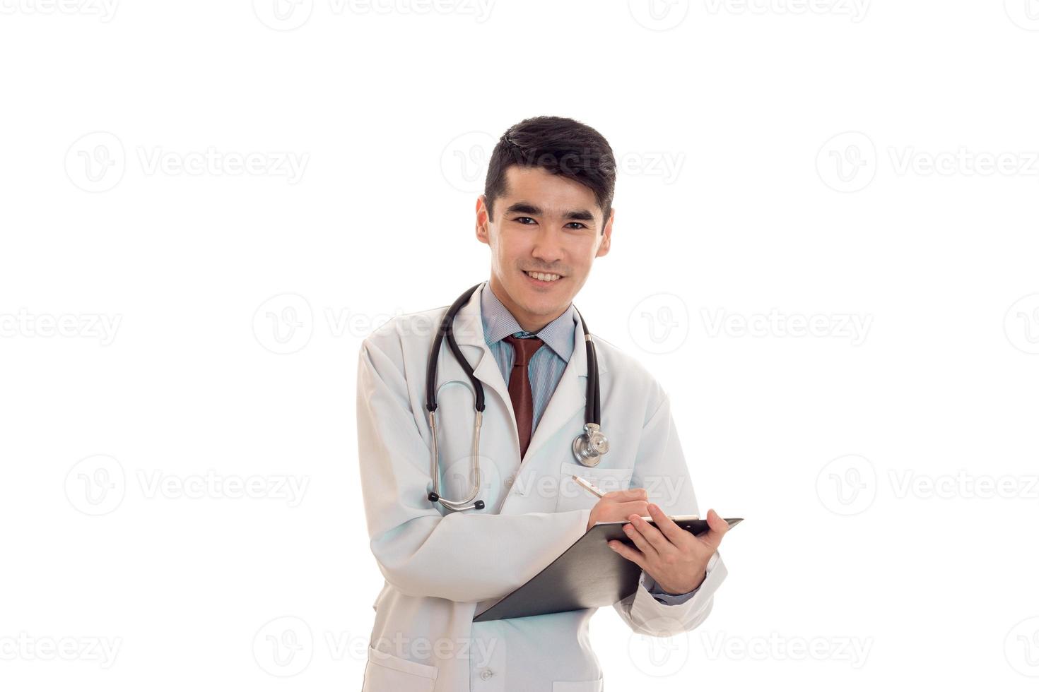 jovem médico alegre mantém um estetoscópio nos ombros e placa na mão isolado no fundo branco foto