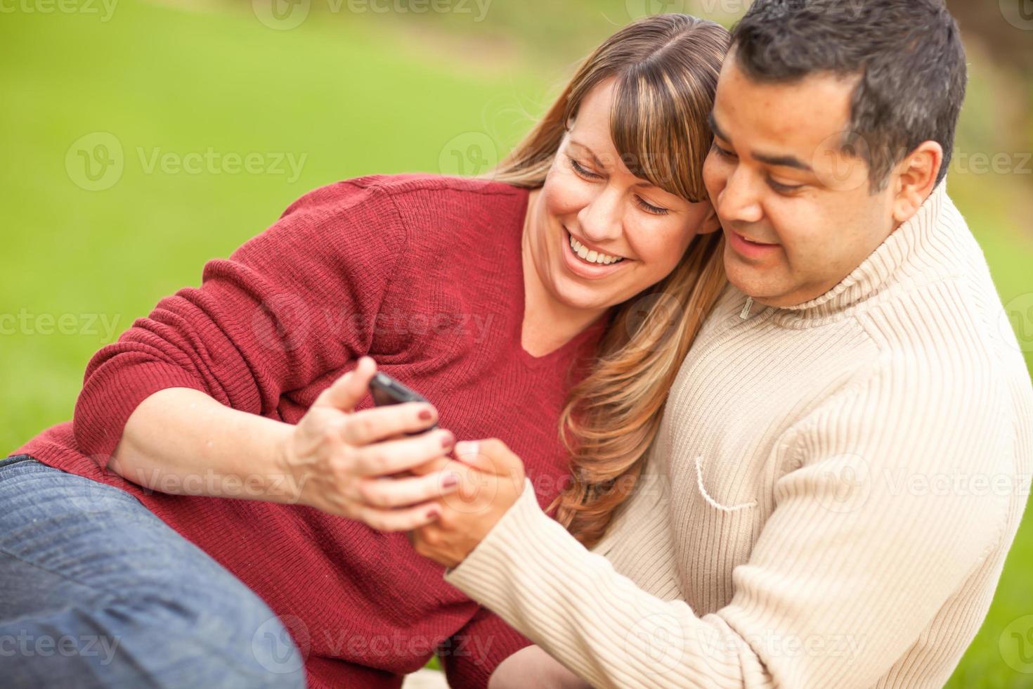 casal atraente de raça mista, desfrutando de seu telefone com câmera no parque foto