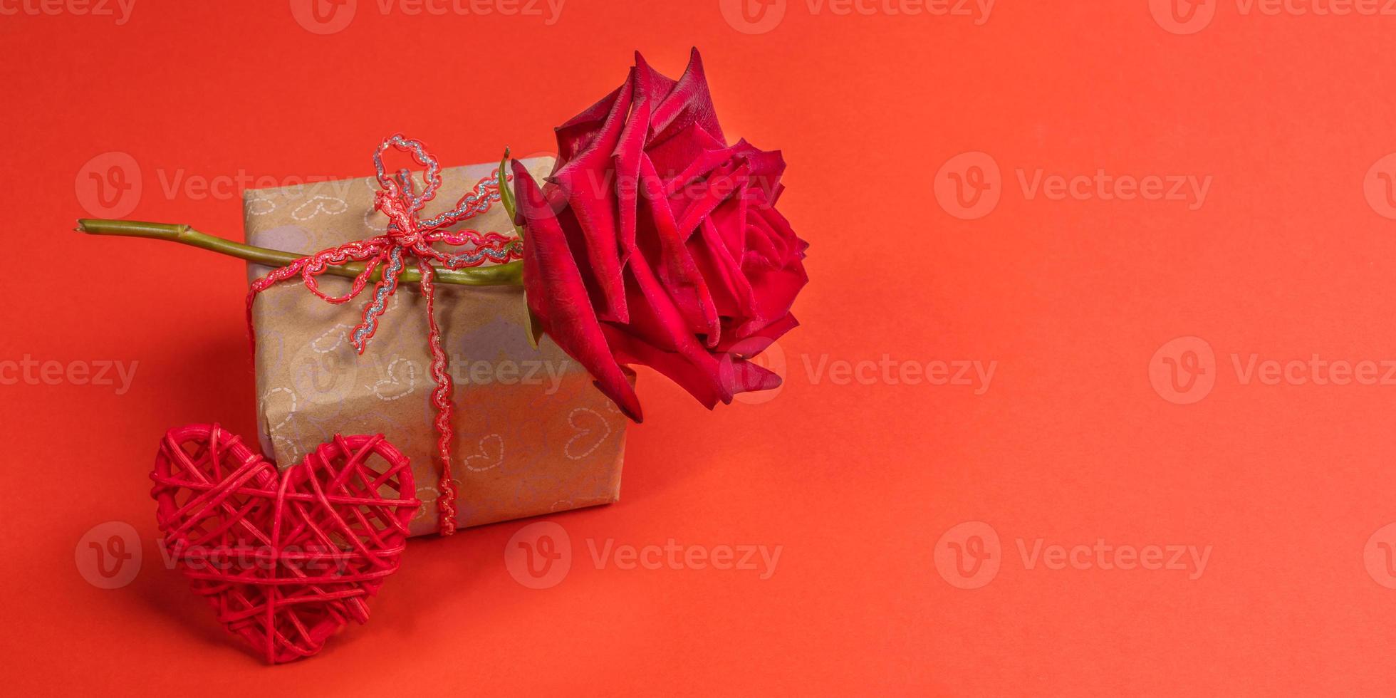 rosa vermelha na caixa de presente embrulhada em papel ofício com corações e coração trançado vermelho sobre fundo vermelho. bandeira do dia dos namorados. foto