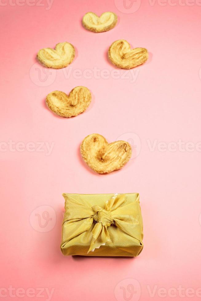 caixa de presente dourada embrulhada na técnica furoshiki e biscoitos em forma de coração em fundo rosa pastel. bolos de férias. foto