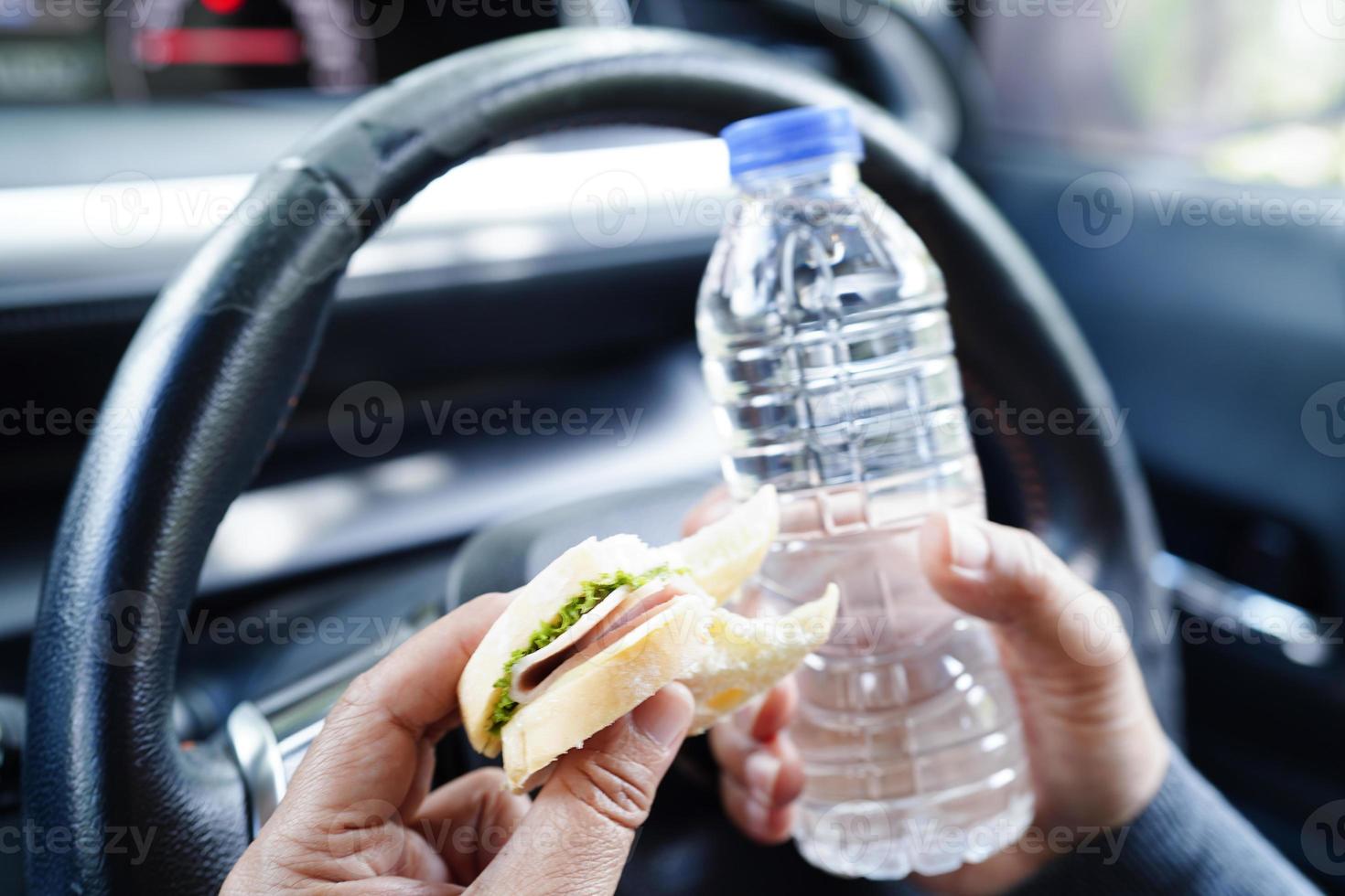 motorista de mulher asiática segura água fria e pão de sanduíche para comer e beber no carro, perigoso e arrisca um acidente. foto