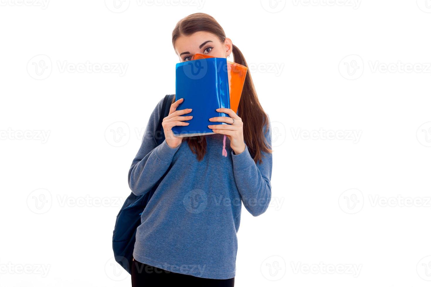 jovem estudante morena engraçada com mochila nos ombros, escondendo o rosto atrás de um livro isolado no fundo branco foto