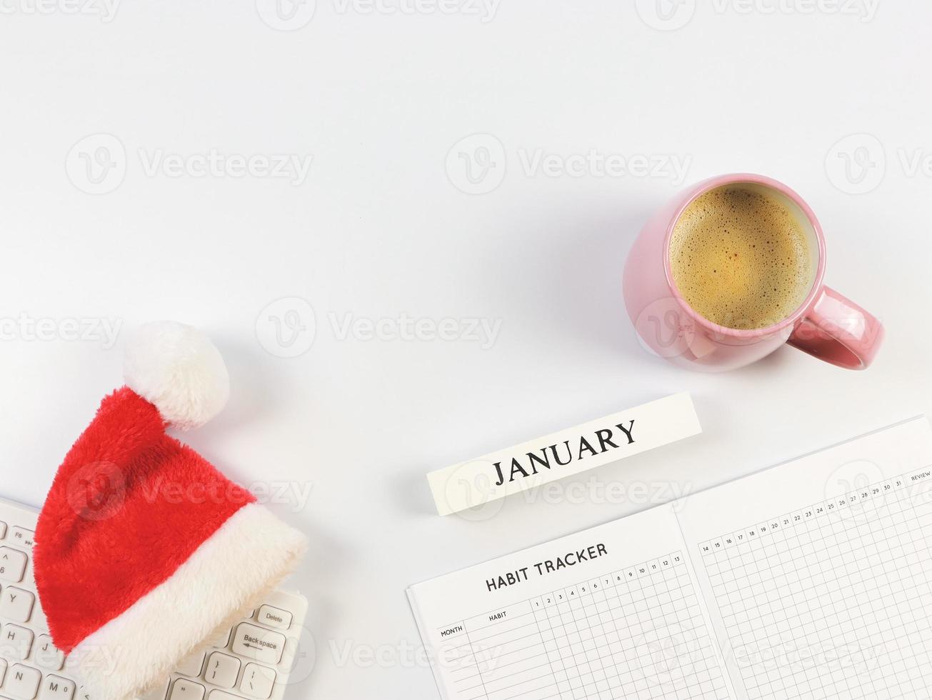 configuração plana do livro rastreador de hábitos, calendário de madeira janeiro, chapéu vermelho de Papai Noel no teclado de computador branco e xícara de café rosa em fundo branco com espaço de cópia. foto