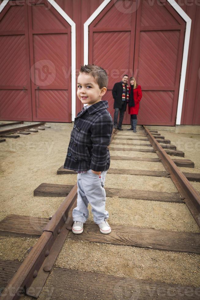 menino de raça mista no depósito de trem com os pais sorrindo atrás foto