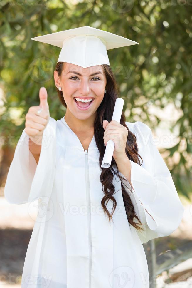 menina de polegares para cima de raça mista comemorando a formatura do lado de fora em boné e vestido com diploma na mão foto