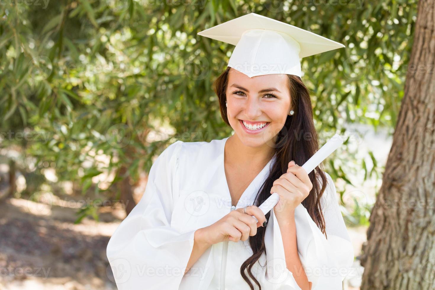 garota atraente de raça mista comemorando a formatura do lado de fora em boné e vestido com diploma na mão foto