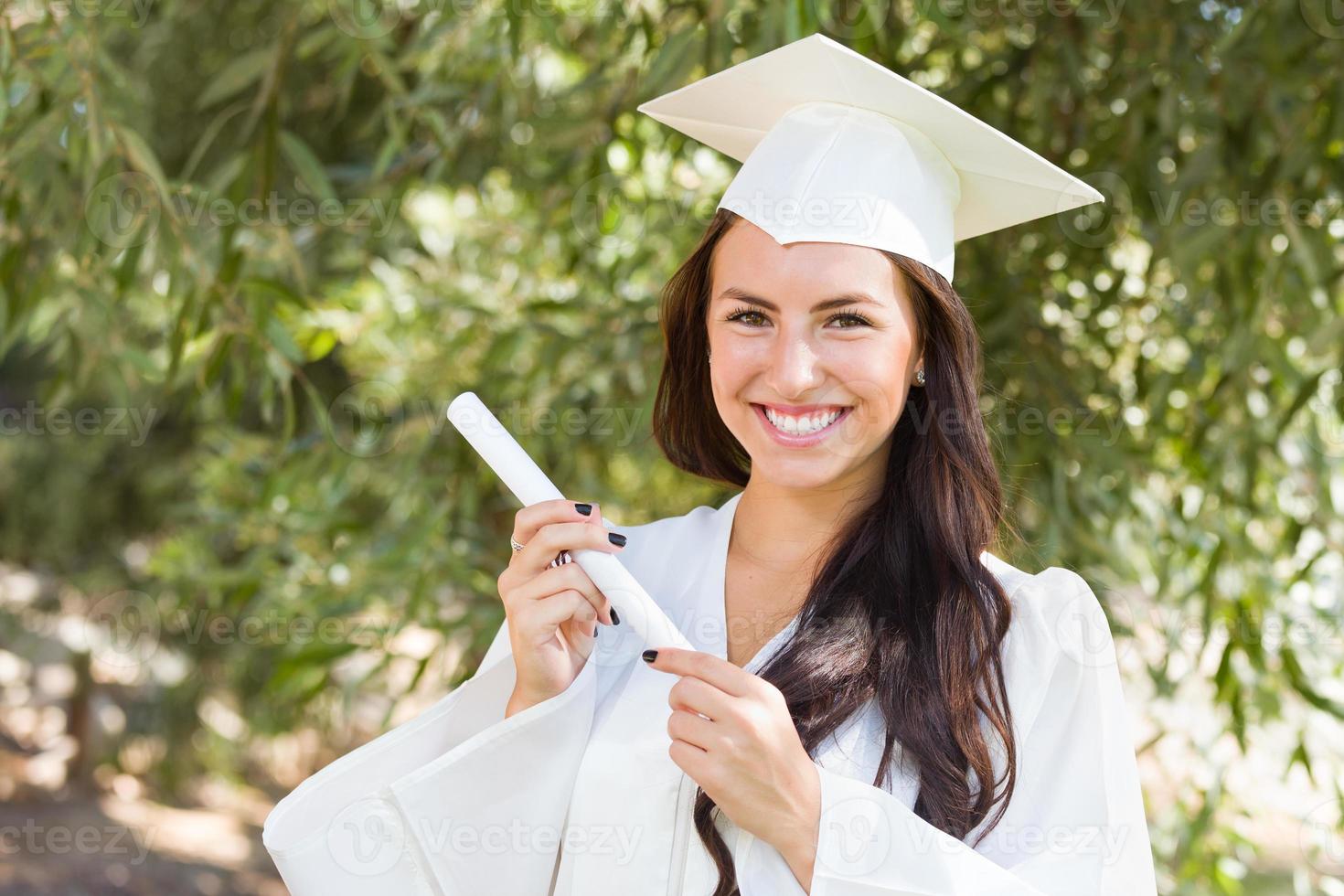 garota atraente de raça mista comemorando a formatura do lado de fora em boné e vestido com diploma na mão foto