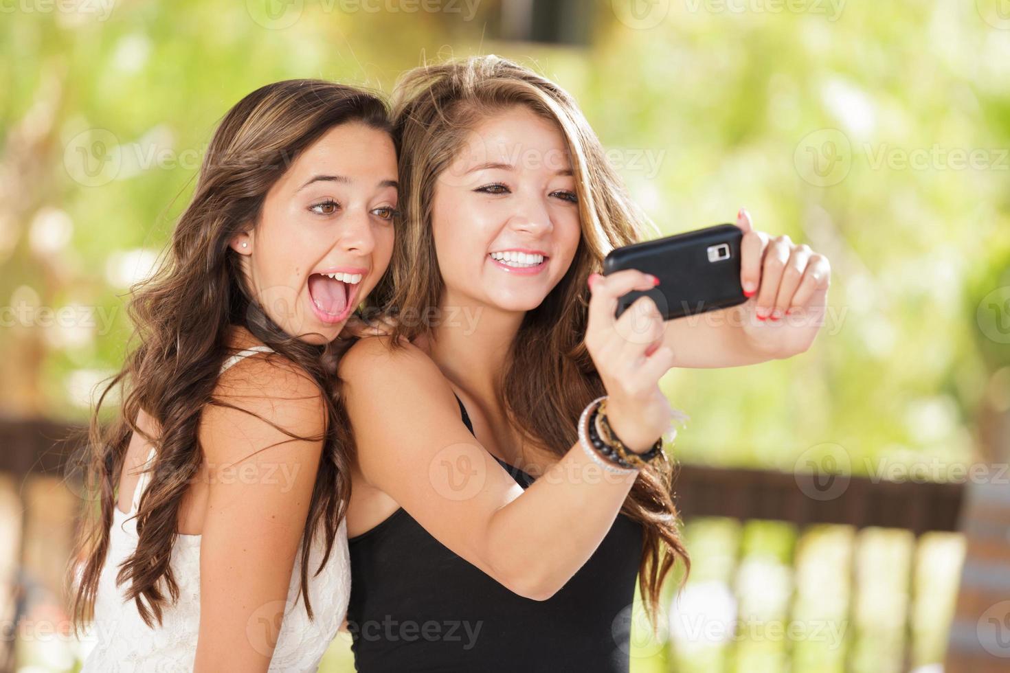 duas namoradas mestiças atraentes usando seus telefones celulares inteligentes para selfie ao ar livre foto