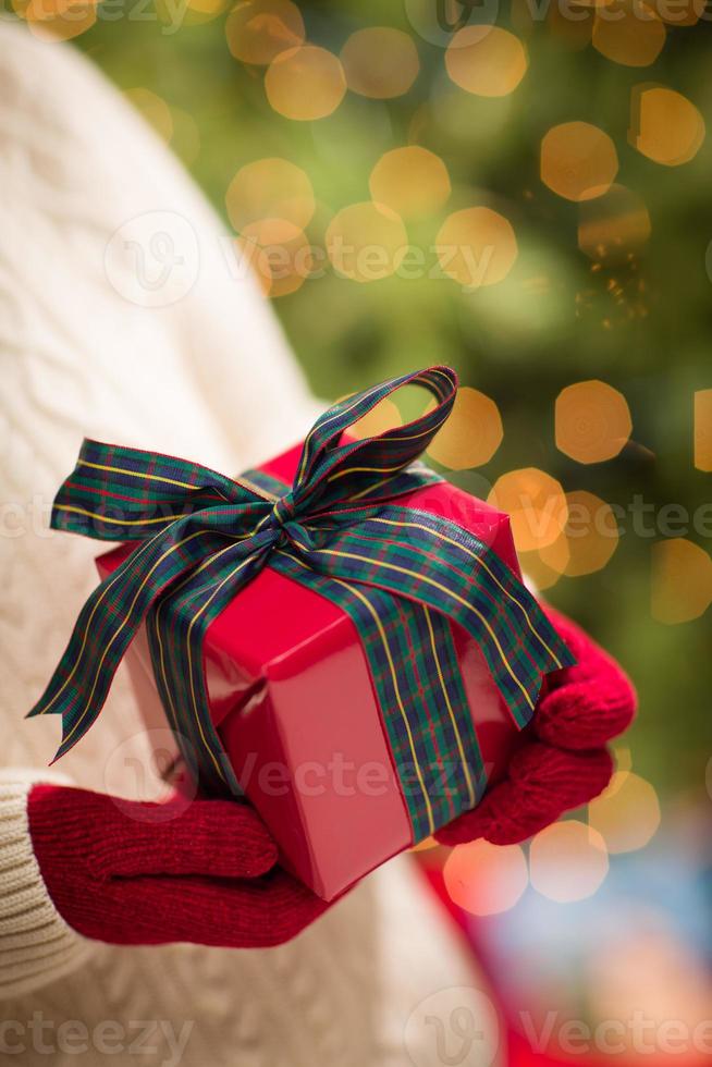 mulher vestindo luvas vermelhas segurando o presente de natal contra a árvore decorada e luzes foto