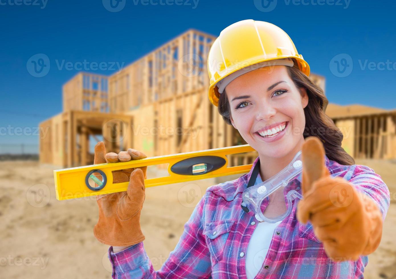 trabalhador da construção civil feminino com polegares para cima segurando nível usando luvas, capacete e óculos de proteção no canteiro de obras foto