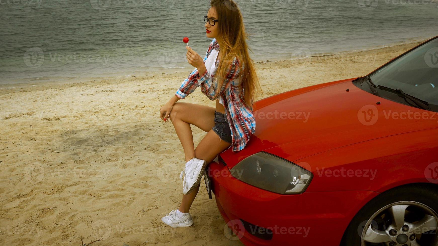 glamour loira de óculos senta-se em um carro vermelho com pirulito nas mãos foto