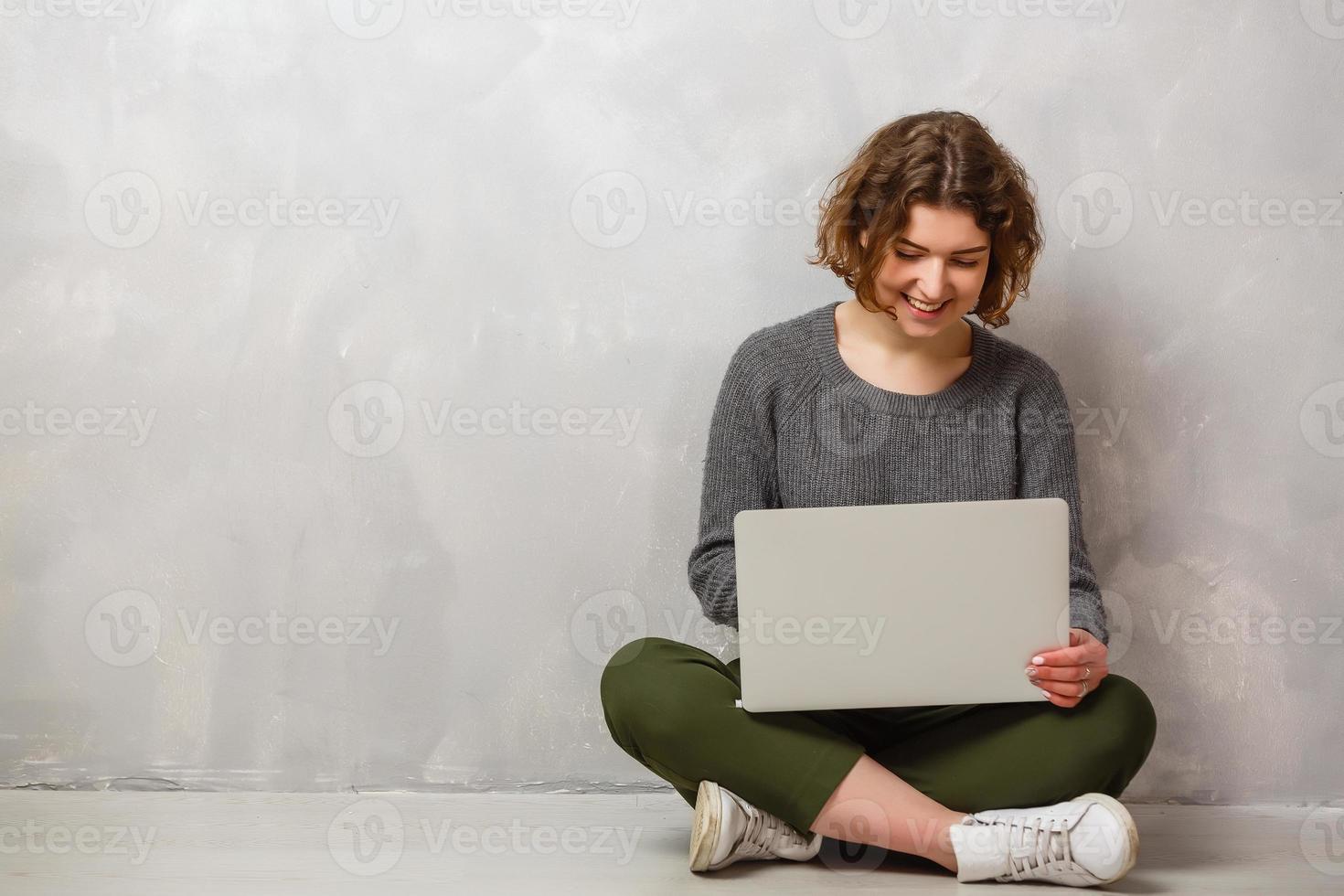 retrato de uma mulher satisfeita com um lindo sorriso desfrutando de assistir filme no computador prateado e sentado em posição de lótus no chão sobre a parede cinza foto