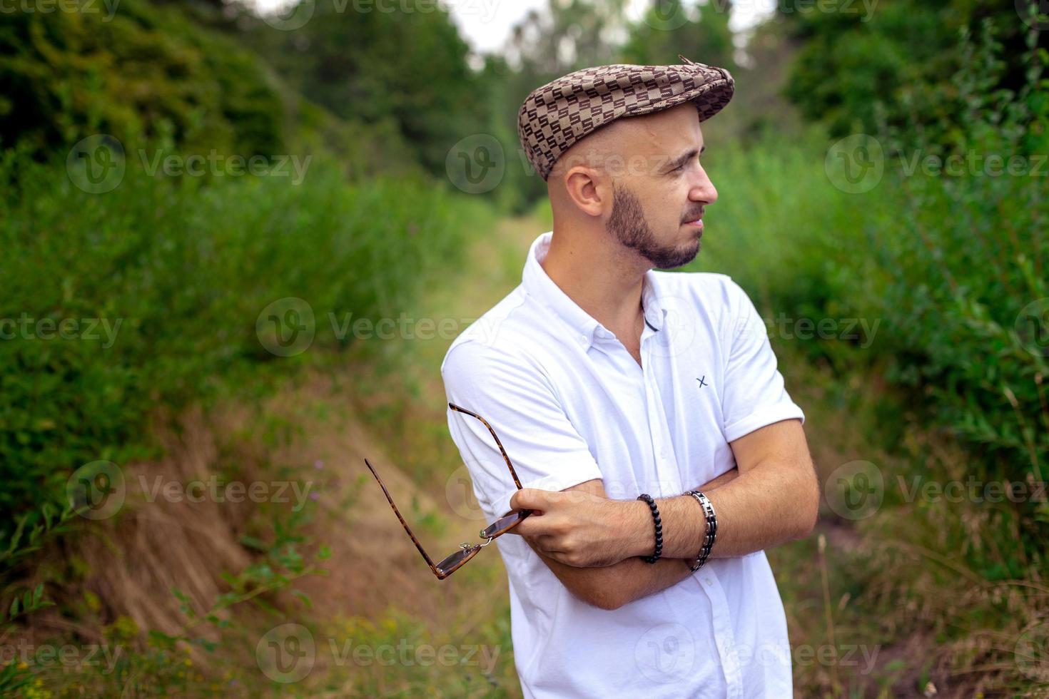 lindo homem de chapéu segura óculos escuros e desvia o olhar para o jardim verde foto