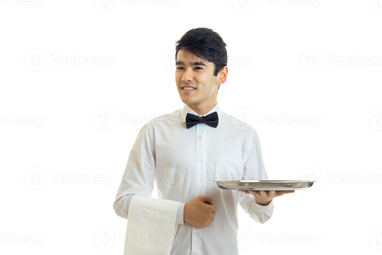 um jovem de camisa branca e gravata borboleta preta desvia o olhar sorrindo e segurando uma toalha e uma bandeja foto