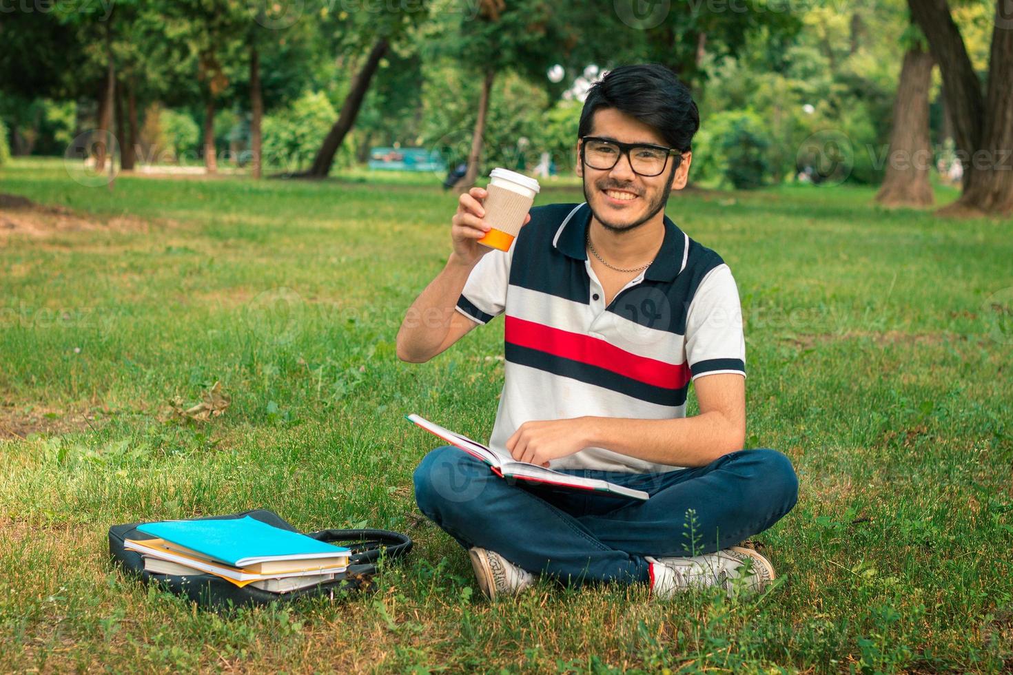 aluno hilário sentado na grama do parque segurando um café e lendo livros foto