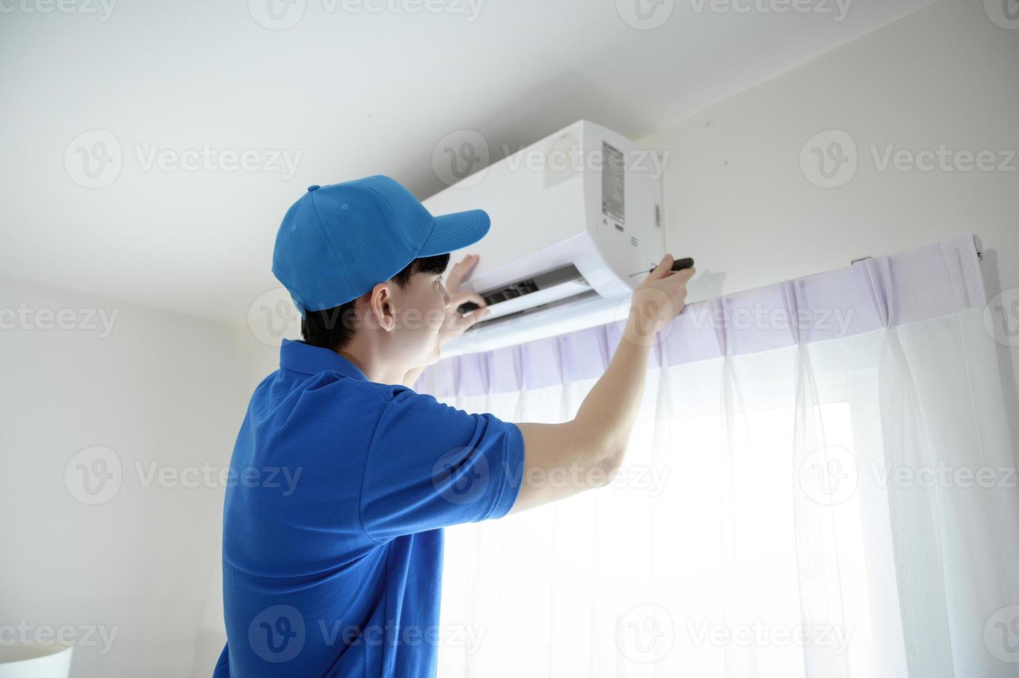 um jovem técnico asiático vestindo uniforme azul verificando, limpando o ar condicionado em casa foto