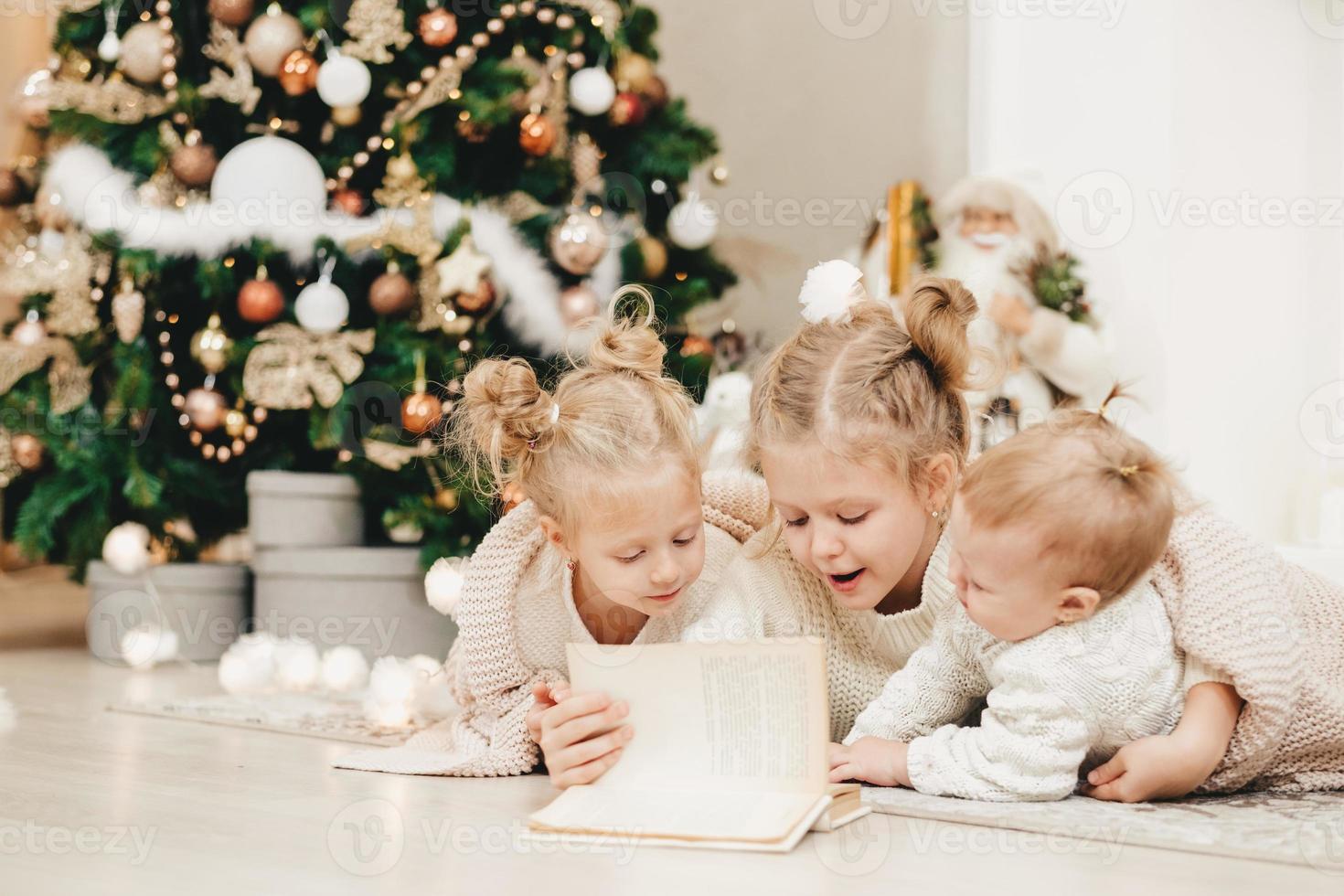 3 garotas loiras deitam no chão perto da árvore de natal e leem um livro. noites aconchegantes com um livro perto da árvore. namoradas leem um livro juntas. relacionamento caloroso foto