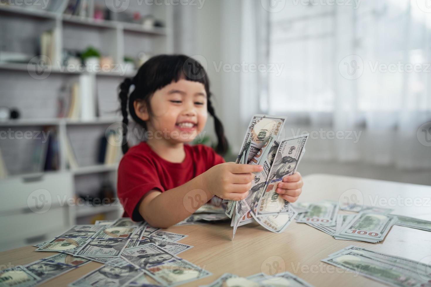 menina asiática vestindo uma camiseta vermelha segurando uma nota de dólar na mesa de madeira na sala de estar em casa. economizando riqueza de investimento, estoque de crescimento, invista juros, fundo, economizando dinheiro para o conceito futuro. foto