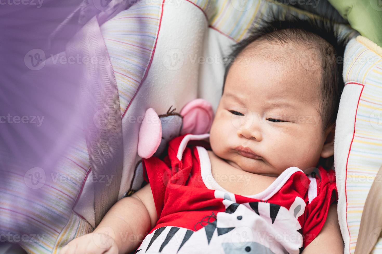 menino asiático de retrato está com sono, a criança está reclinada na cadeirinha. O poder do ventilador roxo embaçado é leve e legal. criança usa macacão vermelho. crianças tem 1 mês, quase 2 meses. foto