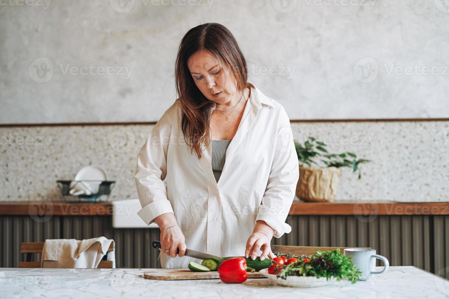 Mulher morena adulta atraente de meia-idade cinquenta anos de camisa branca cozinhando salada de legumes frescos na cozinha em casa foto