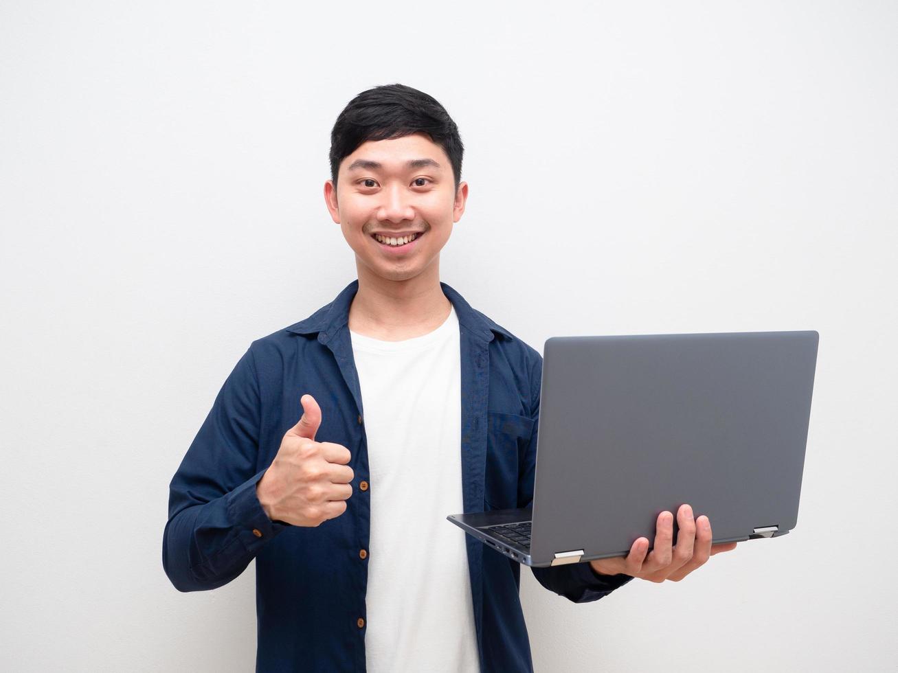 bonito homem asiático camisa azul segurar laptop na mão e polegar para cima com sorriso feliz alegre no fundo branco foto